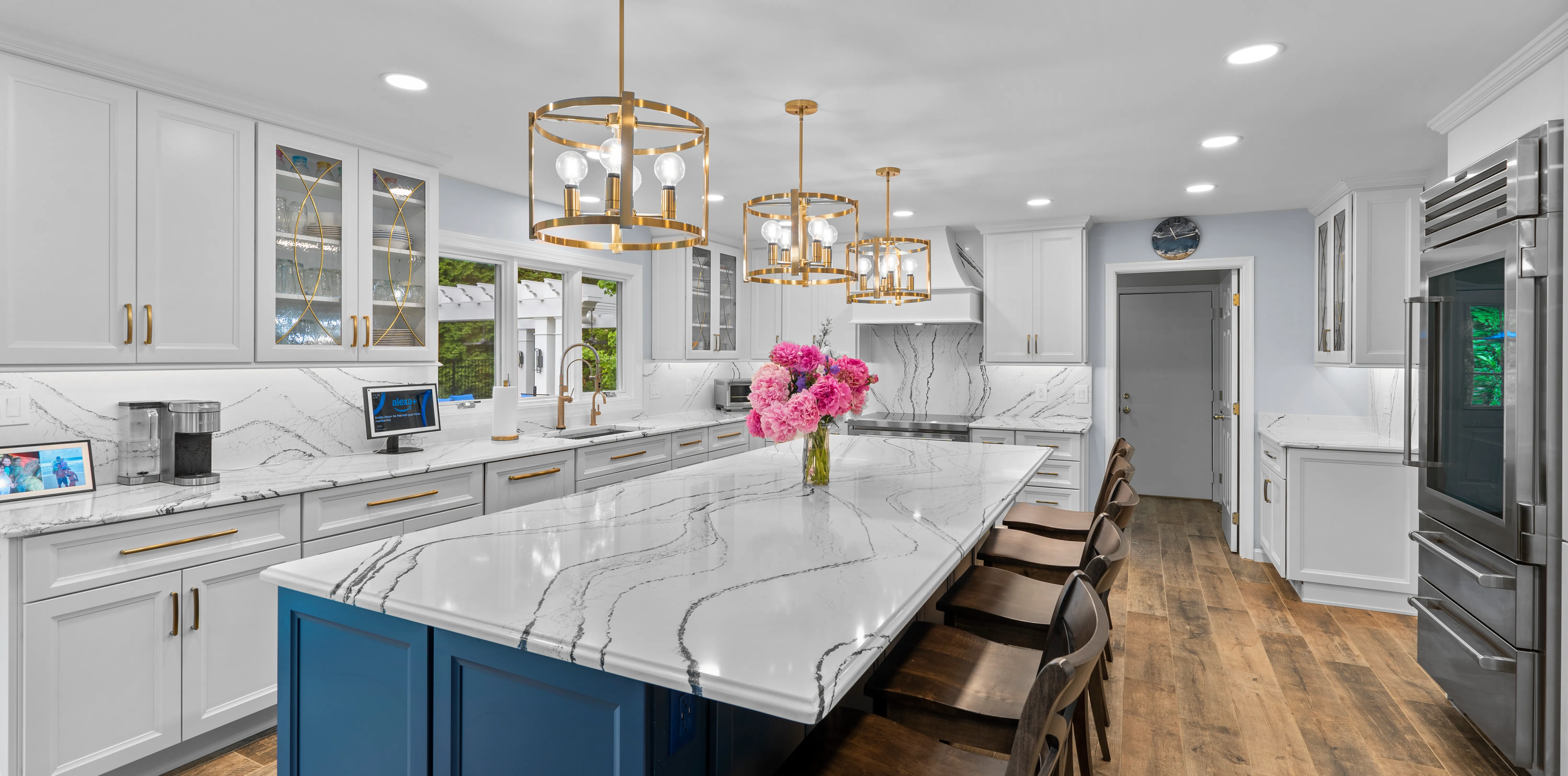 A bright, modern kitchen featuring a large white quartz island with dramatic gray veining, dark blue island cabinetry, gold pendant lights, and white perimeter cabinets with gold hardware remodeled in Gaithersburg, MD by Tile Center.